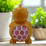 A yellow dinosaur stress toy with a pink mesh gel belly sitting on a white desk with green plants in the blurred background.