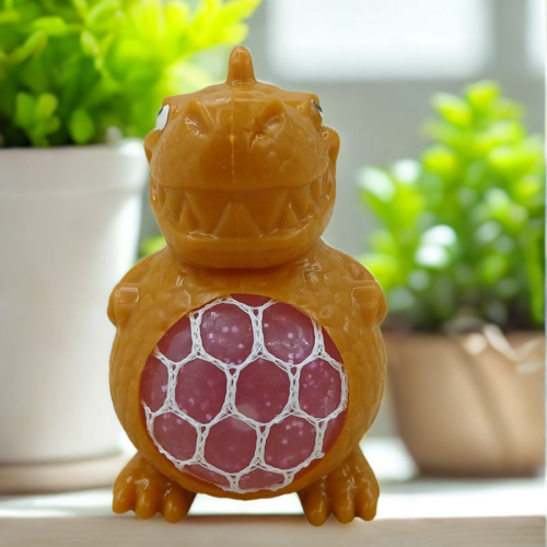 A yellow dinosaur stress toy with a pink mesh gel belly sitting on a white desk with green plants in the blurred background.