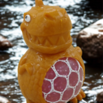 A yellow dinosaur stress toy with a pink mesh gel belly sitting on a white desk with green plants in the blurred background.