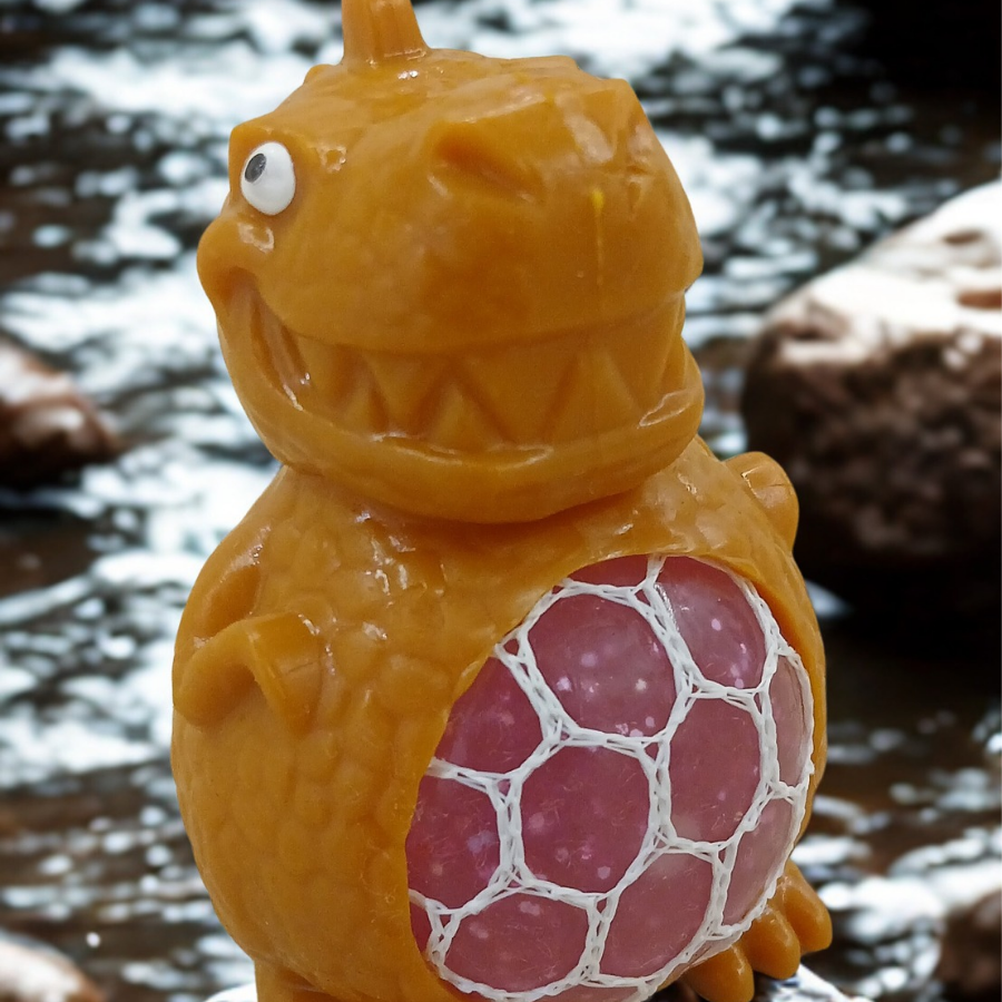 A yellow dinosaur stress toy with a pink mesh gel belly sitting on a white desk with green plants in the blurred background.
