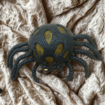 A black spider-shaped stress toy with yellow glitter gel popping through its textured body on a crinkled fabric background.