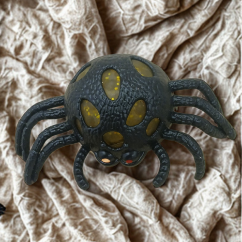 A black spider-shaped stress toy with yellow glitter gel popping through its textured body on a crinkled fabric background.