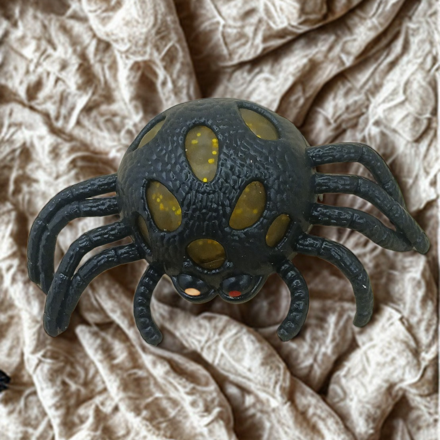 A black spider-shaped stress toy with yellow glitter gel popping through its textured body on a crinkled fabric background.