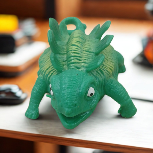 A textured green ankylosaurus dinosaur stress toy with ridges on its back sitting on an office desk setup.