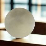 A minimalist, frosted translucent stress sphere resting on a polished wooden desk with a soft-focus office window in the background.