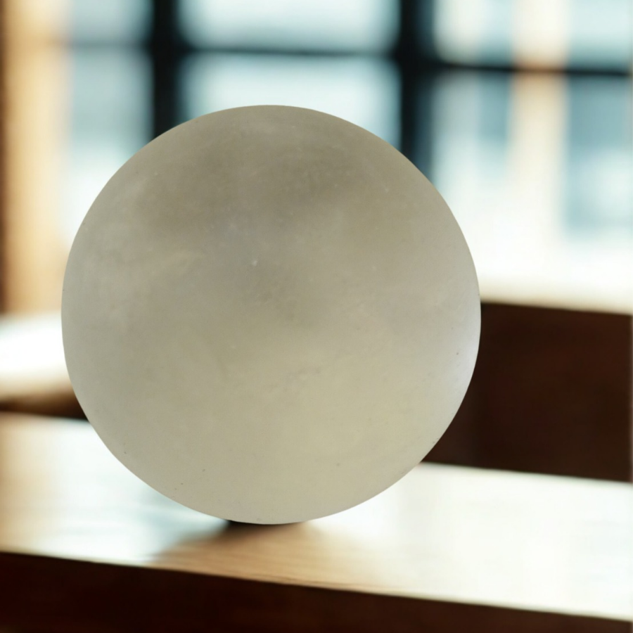 A minimalist, frosted translucent stress sphere resting on a polished wooden desk with a soft-focus office window in the background.