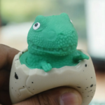 A person squeezing a speckled egg toy to reveal a green dinosaur popping out from the top.