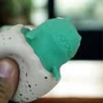 A person squeezing a speckled egg toy to reveal a green dinosaur popping out from the top.