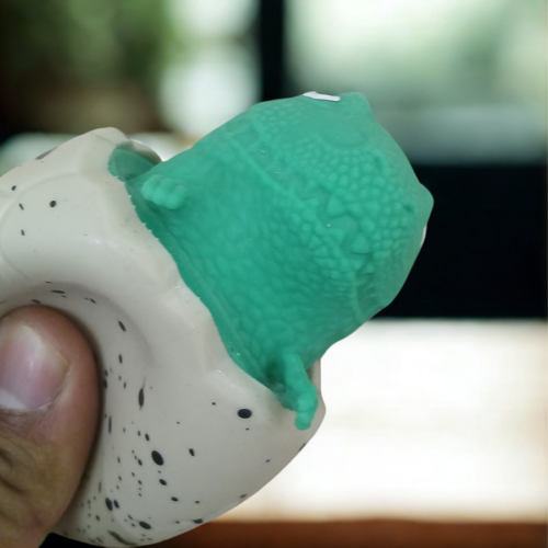 A person squeezing a speckled egg toy to reveal a green dinosaur popping out from the top.